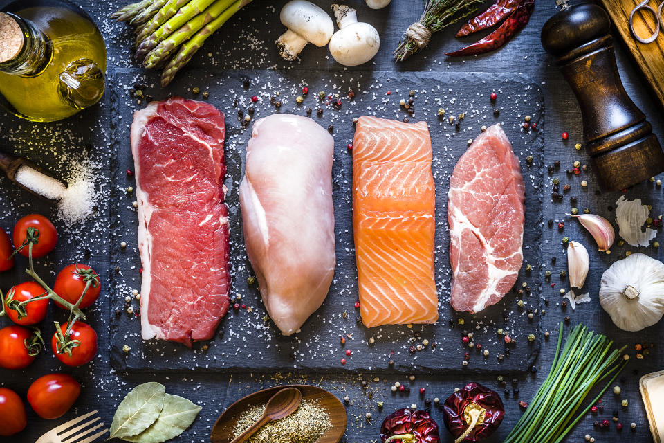 Top view of four different types of animal protein like a raw beef steak, a raw chicken breast, a raw salmon fillet and a raw pork steak on a stone tray. Stone tray is at the center of the image and is surrounded by condiments, spices and vegetables