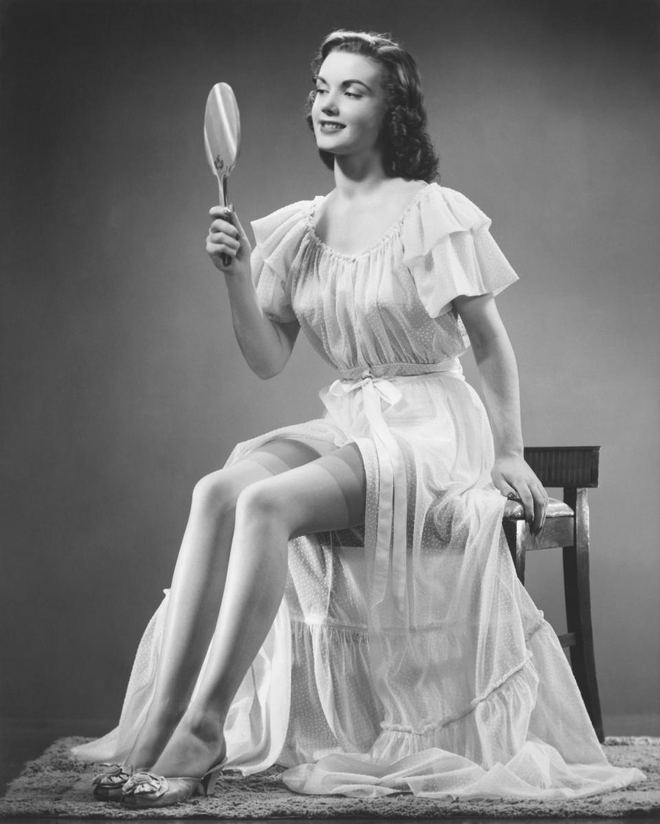 A '30s styled woman perched on a lowback chair wearing a long, flowing nightgown with short sleeves and high-heel slippers, looking into a hand mirror