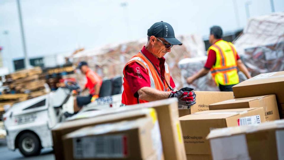 A Delta Air Lines cargo handler handles a box while other employees deal with a sea of cargo.