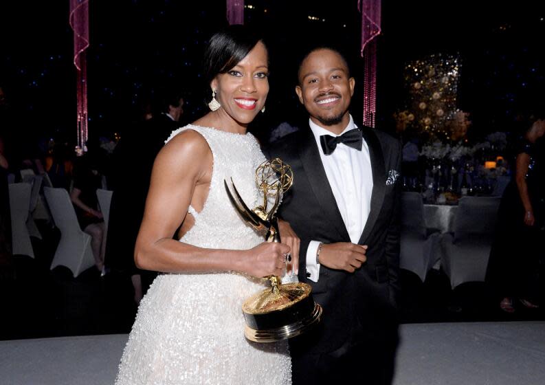 Regina King in a white gown holding an Emmy Award standing next to a young man with a mustache wearing a tuxedo