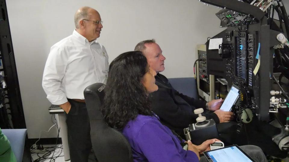 Two people sitting in the simulator look at the screens.  A person stands behind them and watches.