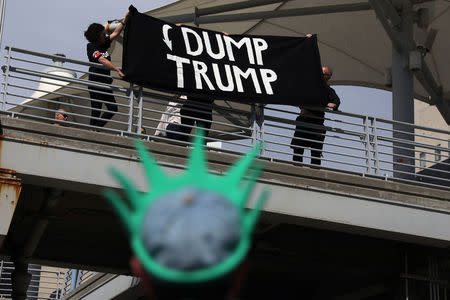 Protesters hold a ""Dump Trump" sign from a bridge to the Intrepid Sea, Air & Space Museum as a person with a State of Liberty visor watches, ahead of an expected visit by U.S. President Donald Trump in the Manhattan borough of New York City, U.S. May 4, 2017. REUTERS/Shannon Stapleton