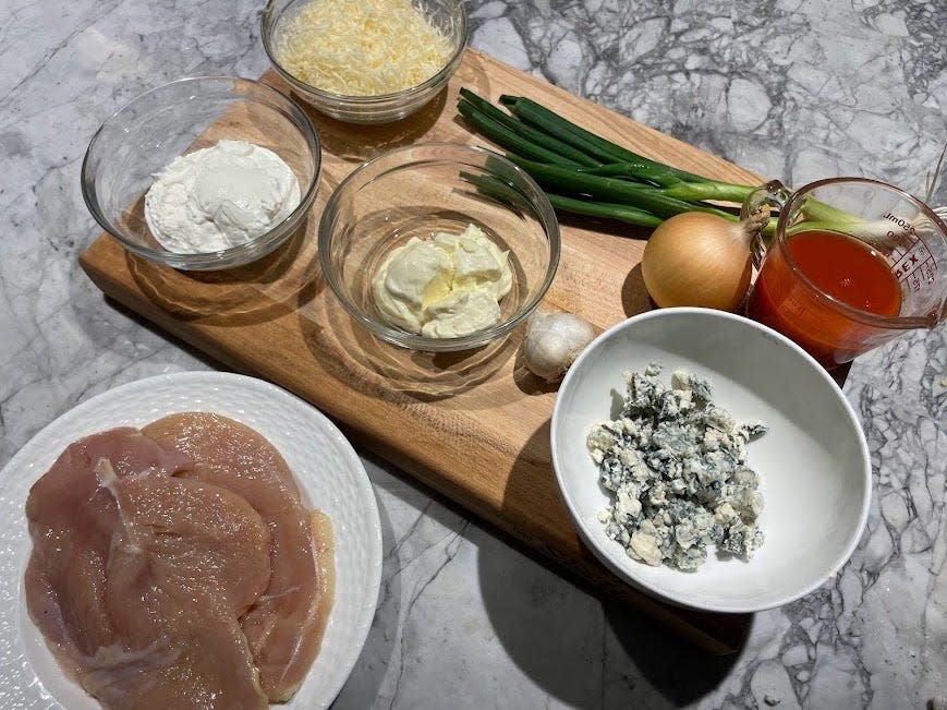 A wooden cutting board with bowls of cheese, sour cream, cheese, a head of garlic, an onion, hot sauce in a measuring cup, and green onions. A plate of raw chicken sits next to the cutting board