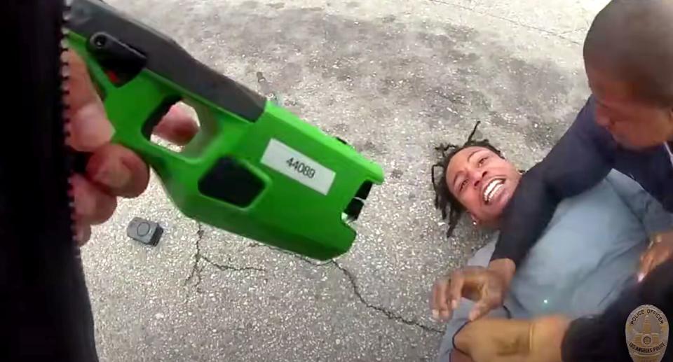 A man is restrained on the ground by a police officer while another officer points a Taser at him.