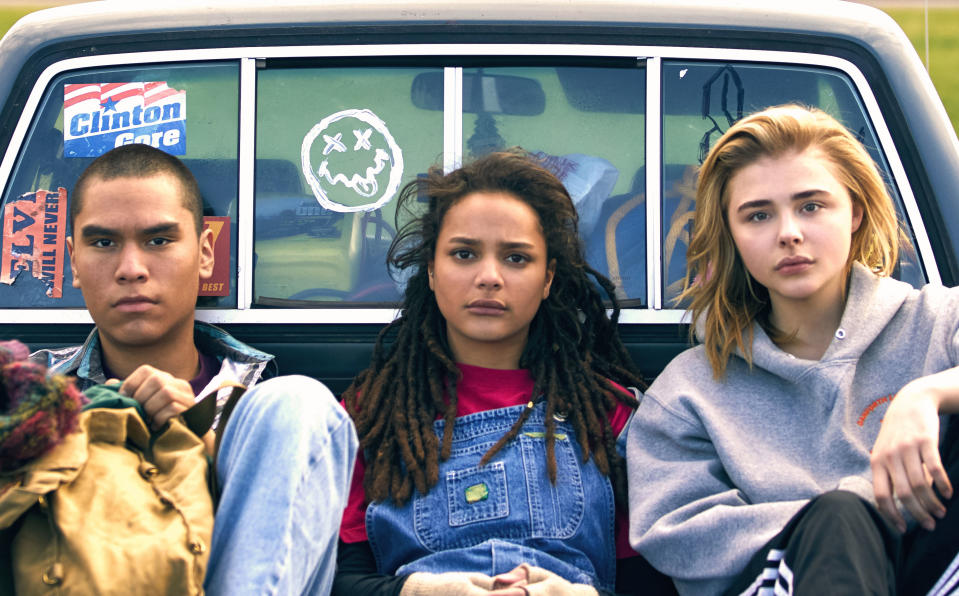 Three people sitting in the back of a truck: Sasha Lane in overalls, Forrest Goodluck in a jacket, and Chloë Grace Moretz in a hoodie. The truck has bumper stickers