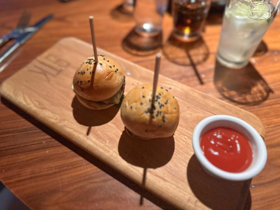 Two small burgers with puffy golden-brown buns, black sesame seeds sprinkled on top, and wooden skewers sticking out of the sandwiches on a wooden serving platter with a cup of ketchup next to it