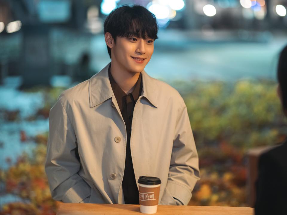 joo yeo-jeong in the glory. he's a kind looking young man smiling while sitting at an outdoor table wearing a grey jacket. there's a hot drink sitting in a travel cup in front of him