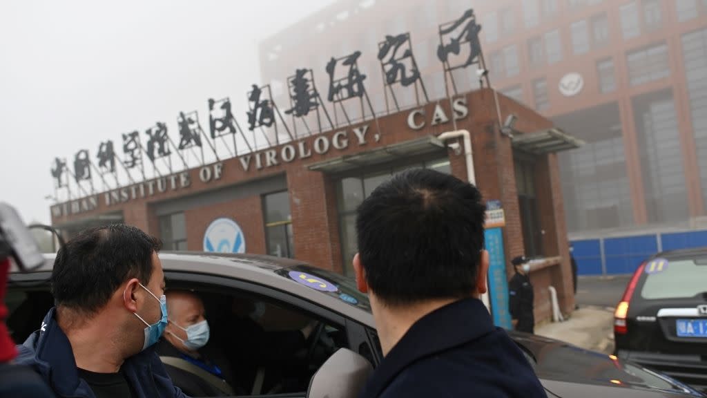  three people in surgical masks, one in a car and two standing nearby, are outside the entrance to the wuhan institute of virology, a multistory building with reddish walls and its name over the door 