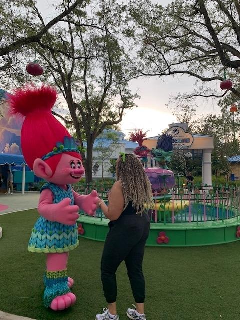 A person interacts with Poppy, a pink character from the Trolls movie, in an amusement park setting. Poppy wears a blue dress