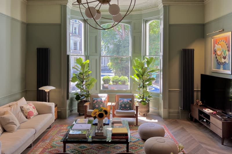 Light green walls with white top trim and bay windows with symmetrical plants and chevron pattern wood floors