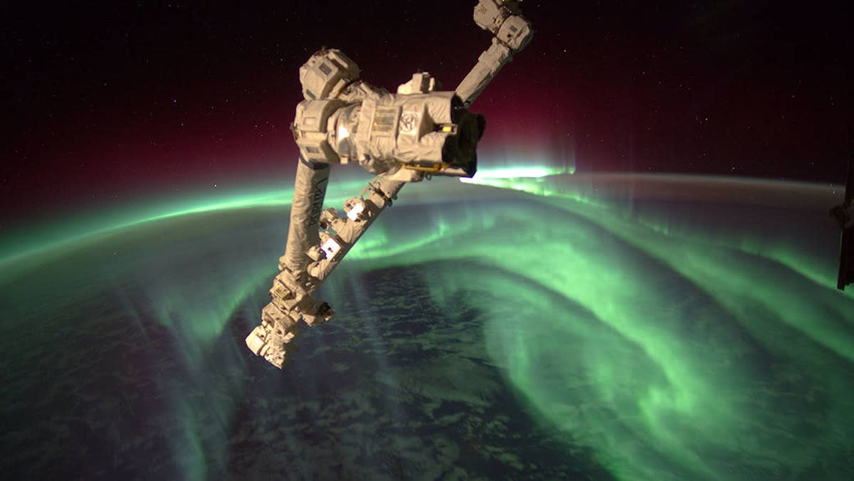  a folded robot arm in front of the earth and black space. the earth is in nighttime and green, swirling auroras are shining above the surface 