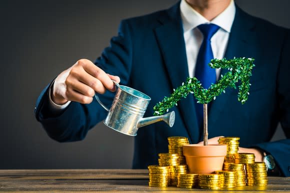 Man in a suit watering a plant shaped like an upward pointing arrow. The plant pot is surrounded by stacks of gold coins.