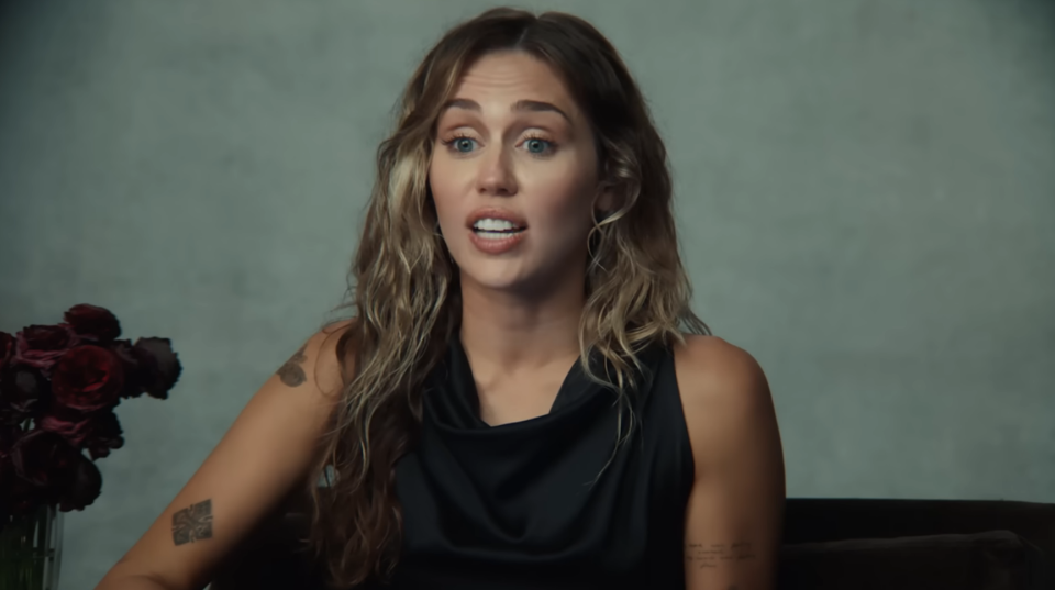 Miley Cyrus speaking in an interview setting with a neutral background and flowers to her left. She wears a sleeveless top