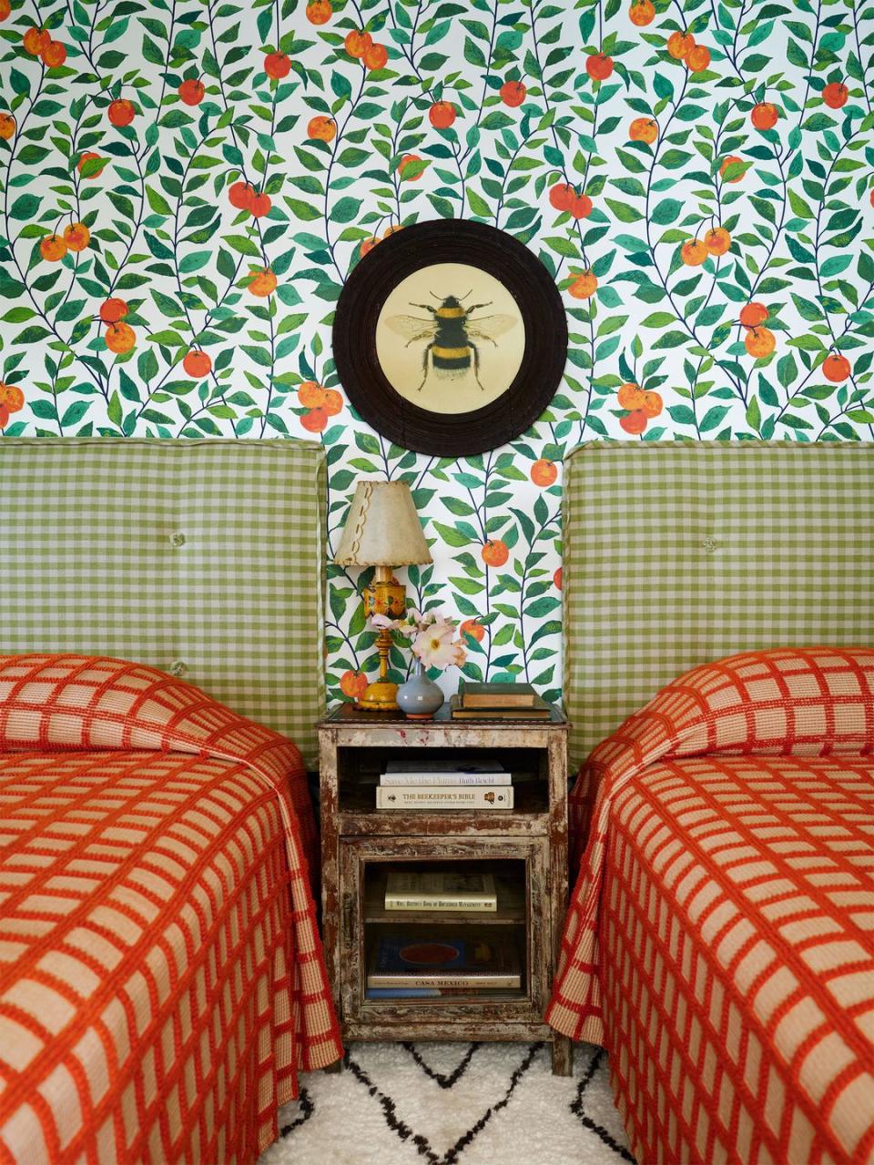 room with wallpaper in oranges with leaves pattern, two beds with an orange reddish popcorn grid spreads, small green check fabric headboards, bumblebee artwork in a round frame, nightstand with lamp