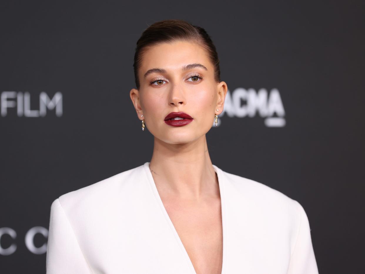 Hailey Bieber at an event in a white dress and dark red lipstick.