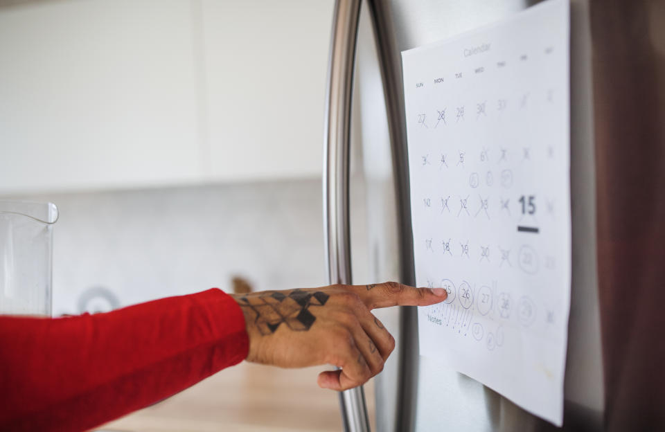 A person's arm with tattooed hand in a red sleeve points to dates on a calendar hanging on a refrigerator, highlighting the 15th of the month