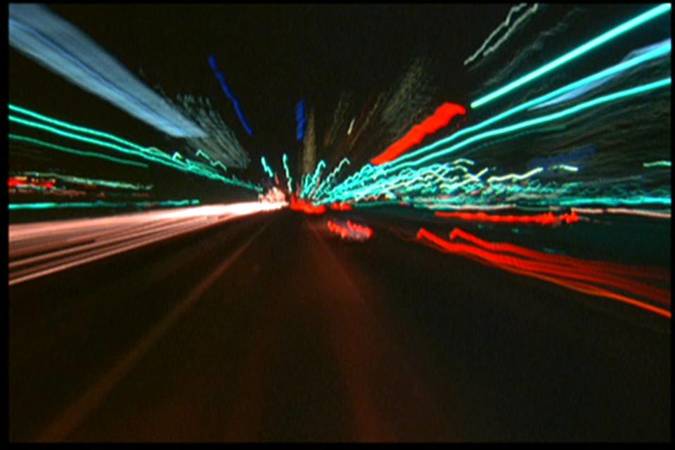 A still from Koyaanisqatsi shows the trails of light from behind traffic