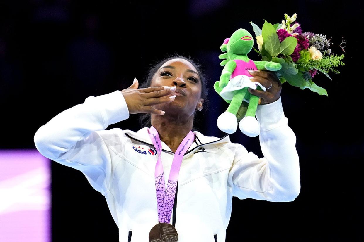 Biles, smiling and wearing her medal, blows a kiss with one hand while holding flowers and a stuffed animal in the other. 