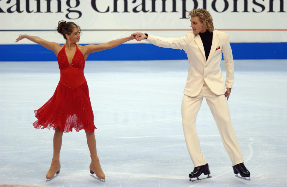 Peter Tchernyshev and Naomi Lang hold hands and face the camera during their competition. She is wearing a red, flowing dress while he is wearing a white suit.