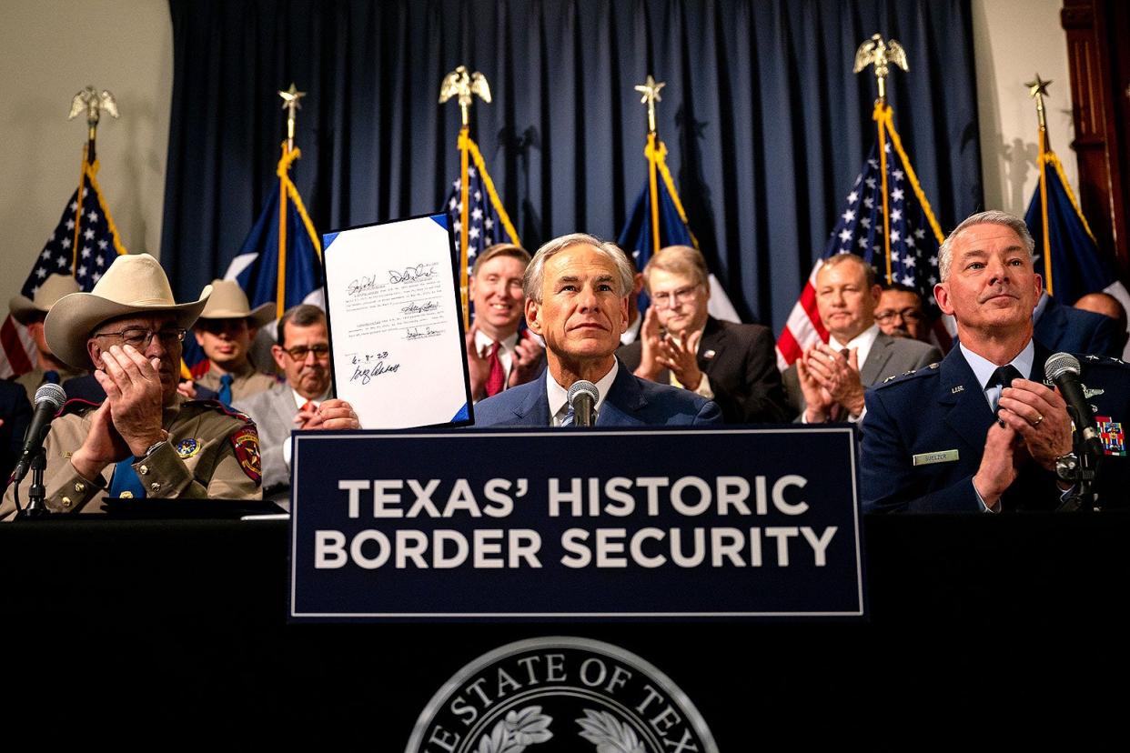 Abbott sits at a table surrounded by officials, some in cowboy hats, holding up a signed piece of paper in front of a podium with the sign "Texas' Historic Border Security."