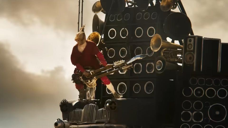 The Doof Warrior in his red pajamas playing his guitar attached to a vehicle with speakers in Furiosa