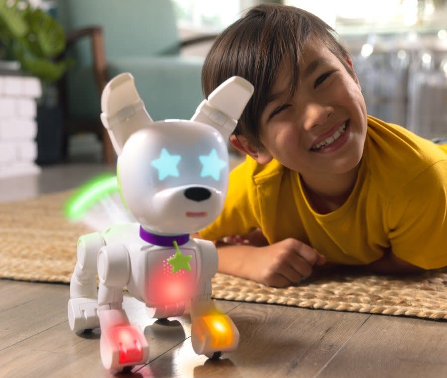 A kid smiles next to a white robotic dog with blue star-shaped eyes