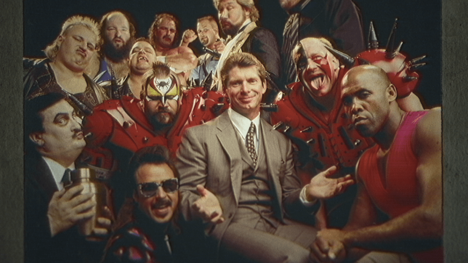 A man in a suit smiles and gestures, surrounded by a group of colorful characters in costumes — wrestlers that worked for his company, including the Road Warriors and their spiked costumes.