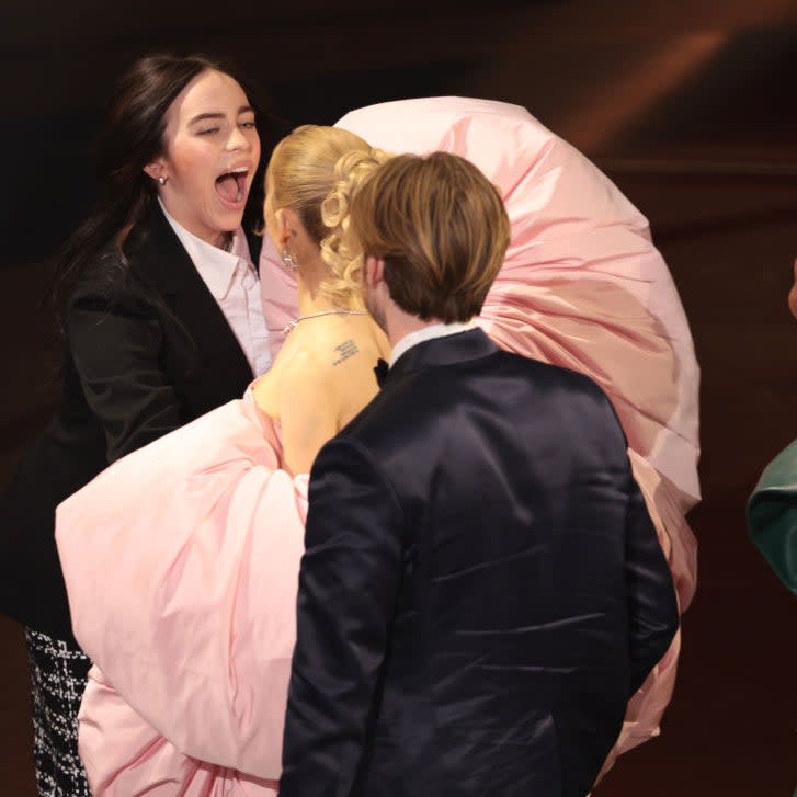 Billie Eilish in a black blazer greets another celebrity wearing a pink dress at an event