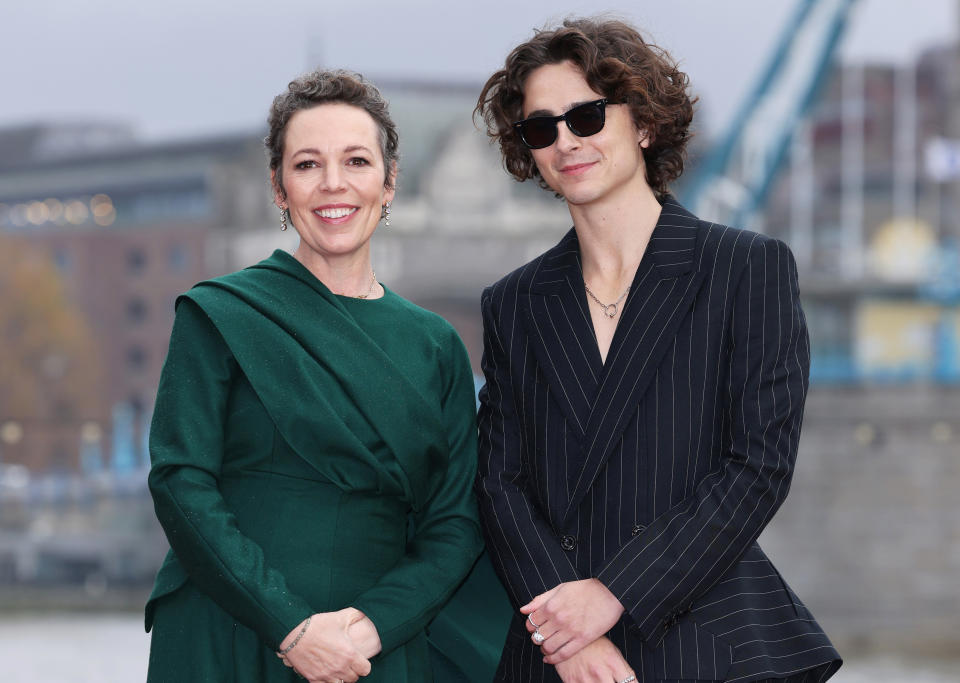 Olivia smiling in a long-sleeved dress with draped detail stands next to Timothée Chalamet, in a pin-striped suit, also smiling