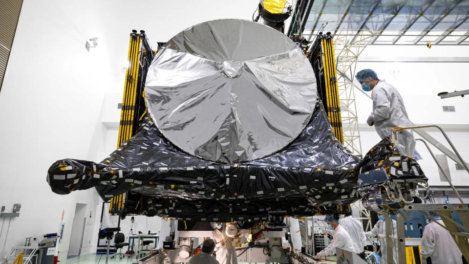 NASA workers in white coats and masks inspecting a spacecraft inside a white building.