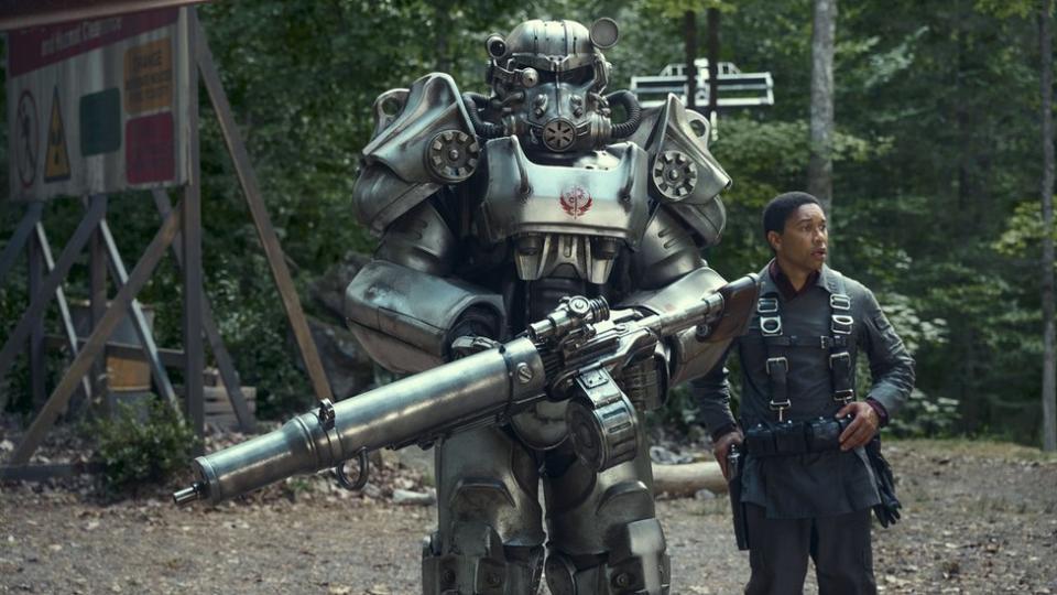 A young black man wearing a black jumpsuit with utility/toolbelt braces stands next to a giant silver figure that towers over him. It's got a robot-like appearance and appears to have been cobbled together from different metallic parts. Covering the face is a large, gas mask style metal covering with an air hose feeding into it. It also holds a large gun with an extended barrel.