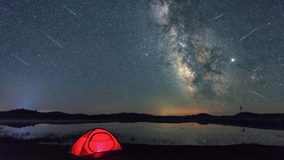 In the sky, streaks of Perseid meteors against the backdrop of the Milky Way and a small red tent in the foreground.