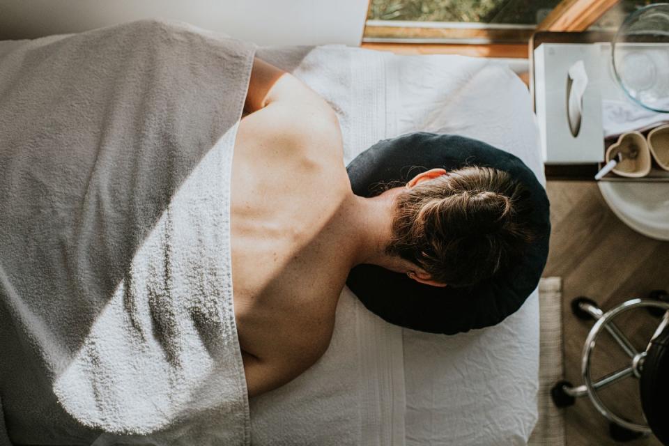 a woman lays face down on a massage table, covered by a towel her bare shoulders and upper back are visible