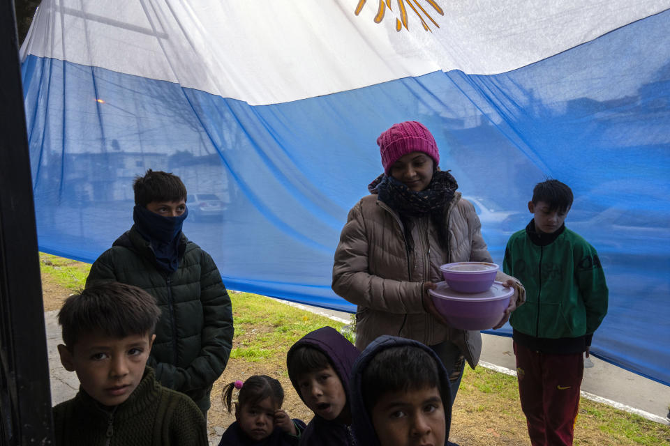 Backdropped by a national flag and surrounded by her children, Celeste Vergara, 29, holds containers of a hominy stew known as Locro, a traditional Independence Day dish that she picked up at the Shinai soup kitchen run by the Movimiento Evita social organization, in Buenos Aires, Argentina, Wednesday, May 25, 2022. Vergara is one of millions of Argentines who survive largely thanks to soup kitchens and state welfare programs, many of which are funneled through politically powerful social movements linked to the ruling party. (AP Photo/Rodrigo Abd)