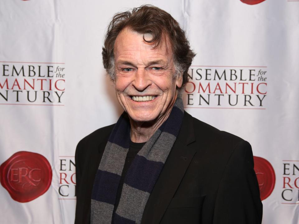 john noble wearing a black suit and gray and blue scarf in front of a white background
