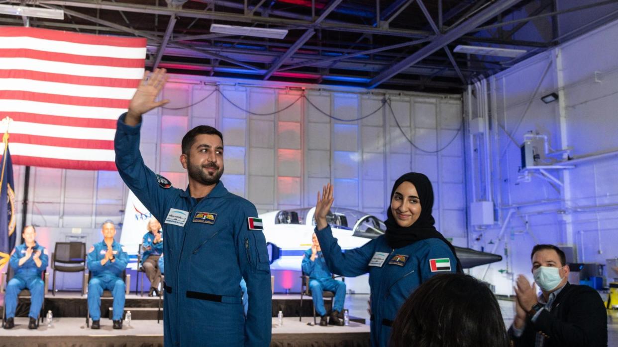  Two astronaut candidates waving in flight suits in front of a us flag and a t-38 jet. on the stage behind, seated astronaut candidates applaud. 