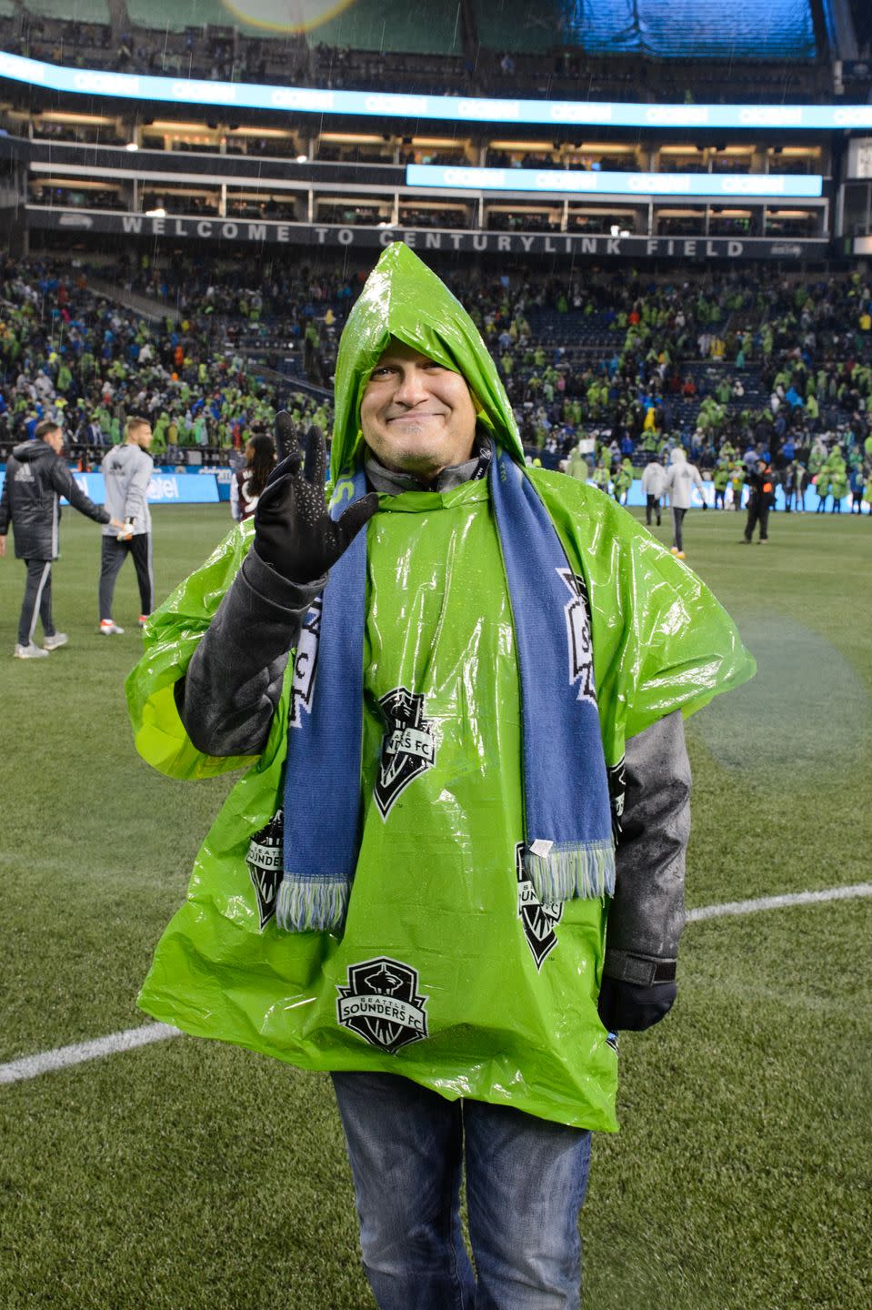 drew carey waving for a photo while wearing a scarf and rain poncho
