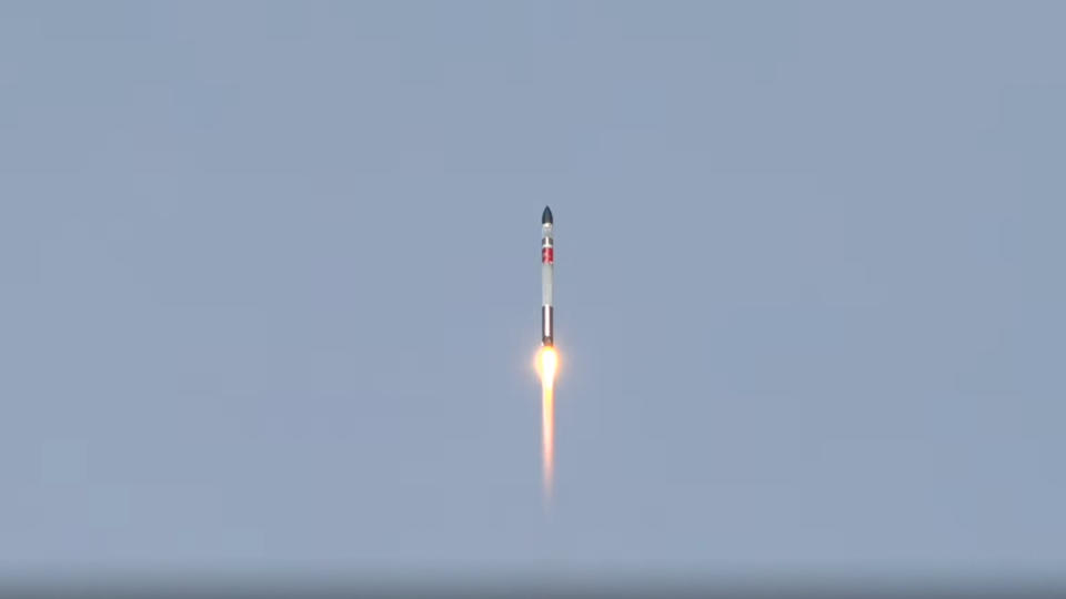 a white, black and red rocket lab electron rocket launches into a blue sky.