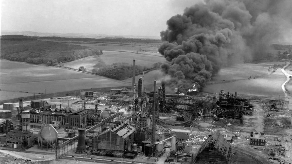 Black and white image showing black plume of smoke emitting from a chemical plant in the middle of a countryside