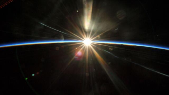 sun rising pierces blackness of space against the dark curvature of the earth with thin blue line of daytime atmosphere on the horizon