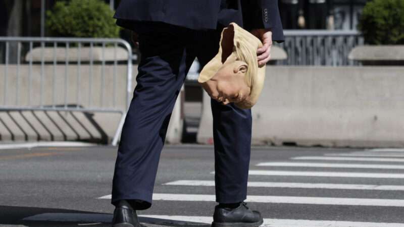 A person crossing the street holding a rubber Donald Trump mask