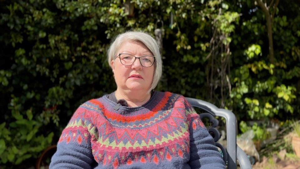 Christine Morgan. She has white hair and is wearing glasses and a jumper while sitting on a chair outdoors
