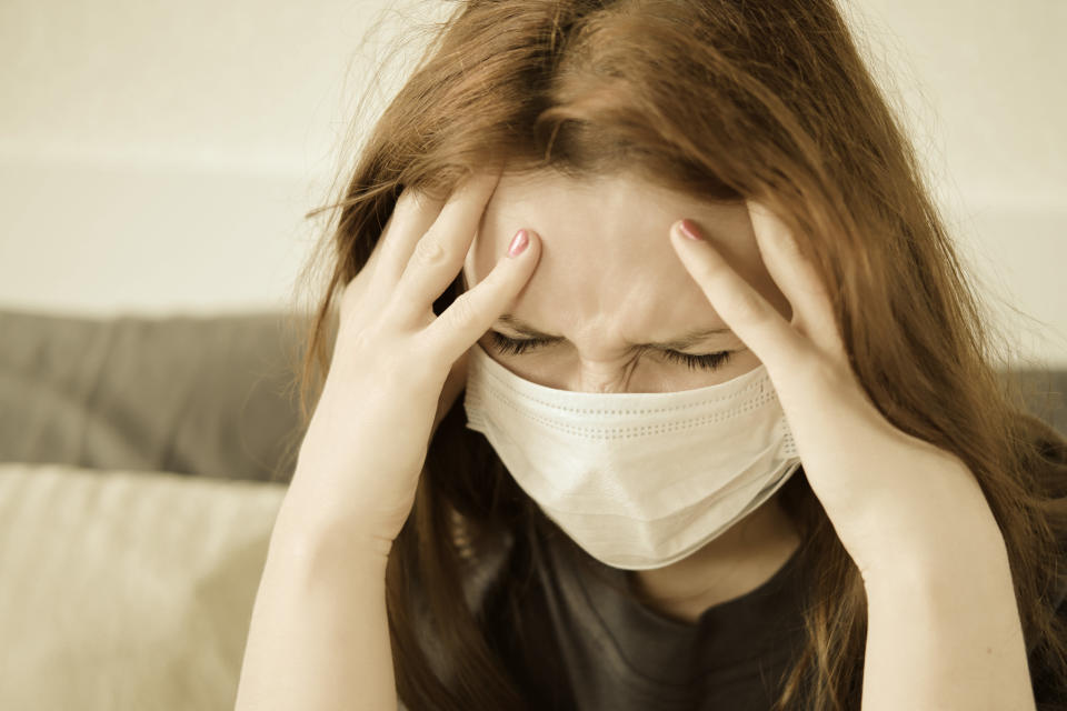 A redhead girl in a medical mask has a headache. Woman with a sore head sits on the bed