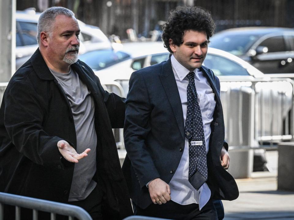 Sam Bankman-Fried, whose shirt is untucked and tie twisted after getting through the media scrum, is escorted by a man in gray shirt and black jacket.