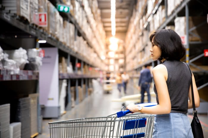 A dark haired woman wheels an empty shopping cart down an aisle of a large warehouse store looking at items.