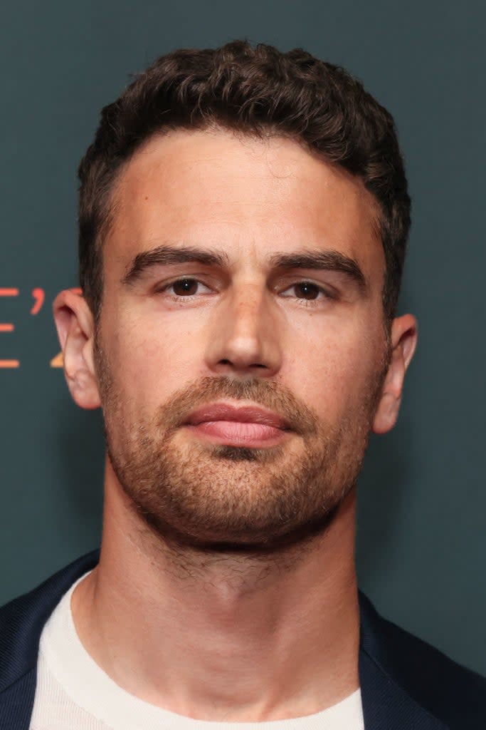 Theo James poses for a photo at an event, wearing a white shirt with a dark jacket