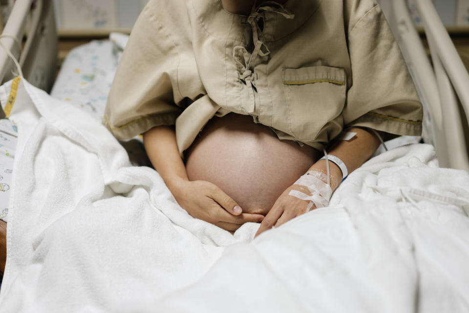 Pregnant woman in a hospital gown sitting on a hospital bed with an IV in her arm, holding her belly