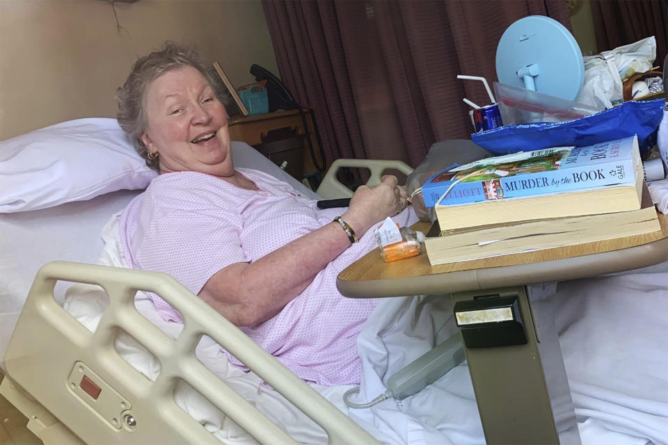 In this March 29, 2020, photo provided by Courtney Templeton, her mother, Sharon Templeton, who tested positive for COVID-19, smiles through her room window at Sundale nursing home in Morgantown, W.Va. Courtney Templeton faults the home for not testing residents fast enough and not keeping healthy ones separate from those just back from a nearby hospital showing COVID-19 symptoms, including her mother’s roommate. (Courtney Templeton via AP)