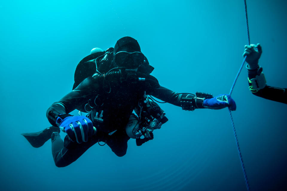 A scuba diver, fully equipped with diving gear and holding a rope, is underwater. Another person's hand, also holding the rope, is partially visible