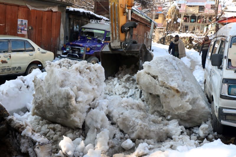 Residents stand as an excavator removes the snow boulders on a road after a heavy snowfall in Neelum Valley